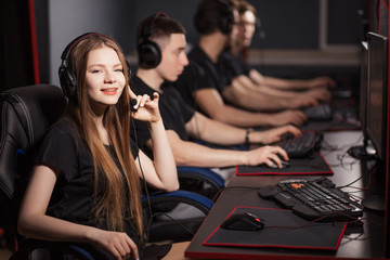 Obraz na płótnie Canvas A cute female gamer girl with headset smiling at camera sits in a dark room behind a computer with a raw of another gamers sitting in line beside her in computer club.