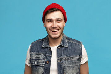 Positiveness, joy and happiness concept. Picture of unshaven friendly joyful young European man in red headwear and jeans vest smiling broadly being in good mood enjoying carefree time of his youth