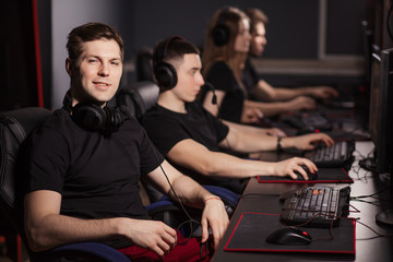Fototapeta na wymiar Satisfied smart gamer in black t-shirt sitting behind the computer monitor and looking at camera in a gaming club or internet cafe with the other people playing.