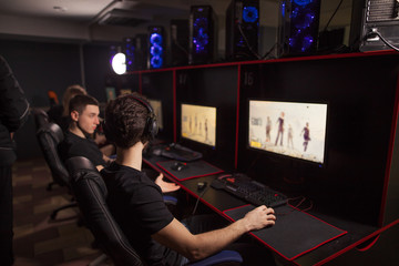 Rear view of focused young gamers sitting in line behind monitors in headsets, playing competitive online game at dark pc gaming club, © alfa27