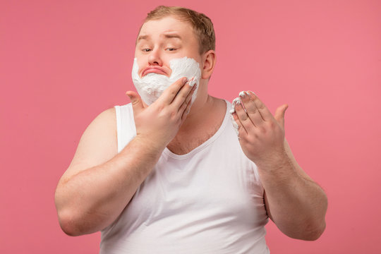 Morning man with foam on cheeks, has sad displeased expression, tired of shaving every morning dressed in white undershirt , isolated over pink background