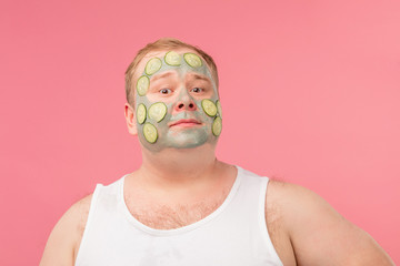 Happy funny tubby man with facial clay mask for skin care having fun with cucumber slices isolated over pink background