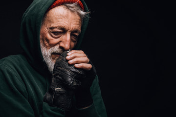 Face portrait of gloomy experienced grey-haired old-aged man with skin lined with wrinkles, dressed in warm shabby clothing as a tramp, rubbing his cold hands with hopeless humble expression.