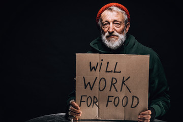 Jobless poor old aged male beggar in orange warm hat looking at camera with a handwritten sign for...