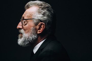 Profile headshot of intelligent bearded grey haired scientist listening report, dressed in formal black suit, sitting isolated over dark