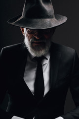 Old-aged bearded man in image of English secret agent wearing black suit with hat on his eyes posing against dark background.