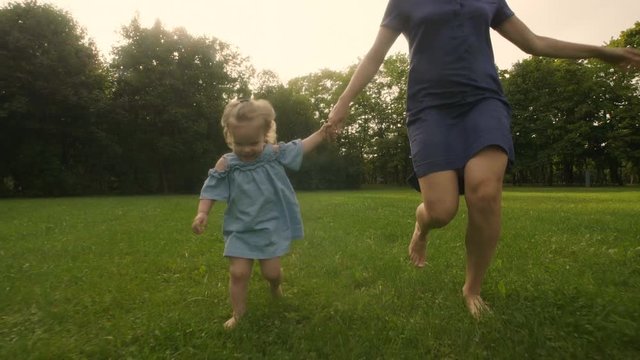 Baby girl with mother running through the grass in the summer sunlight in park. Baby have fun. Slow motion. Happy family concept. 