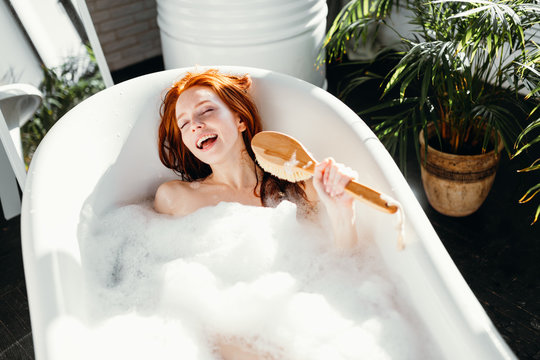 Vivacious emotional young woman with red hair bun taking bath at home, being in good temper, singing a song holding body brush as a microphone, giving way to her talant and joy