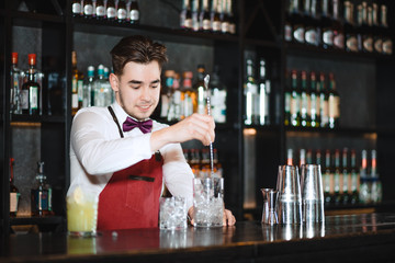 Barman holding a long spoon and glass filled with ice cubes on the bar counter of night club