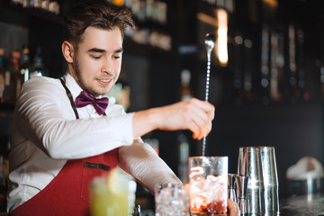 Barman holding a long spoon and glass filled with ice cubes and red drink mixing the beverage on the bar counter of night club