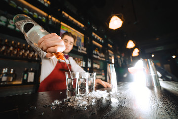 Low angle shot of bartender pouring some drink from the bottle into shot frosted glasses with...