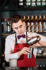 Young handsome male bartender is mixing ingredients in shaker and pouring alcohol from shaker to glass at bar background. Barman at work.