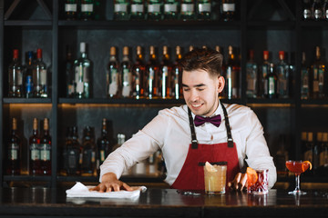 Professional young bartender in white shirt and red apron, wearing bow tie posing at work in night club at bar stand. Service and Entertainment business.