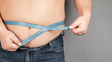 A man measures his fat belly with a measuring tape. on a gray background.