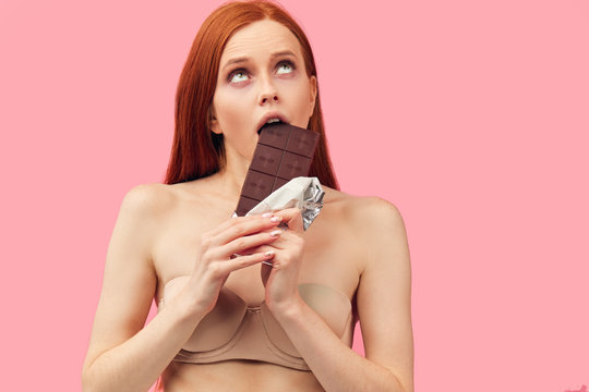 Young skinny anorexic unhealthy woman is fighting with temptation to eat chocolate bar, looking looking miserable at camera, isolated over pink background