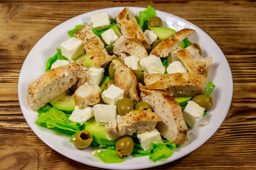 Tasty salad with fried chicken breast, green olives, feta cheese, avocado, lettuce and olive oil on wooden table