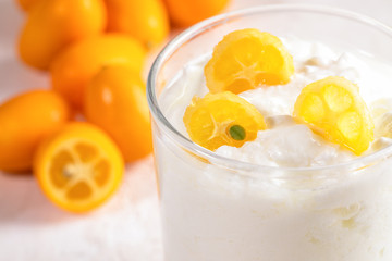 Close-up part of glass cup with yogurt with slices of kumquat on whole fruits background.