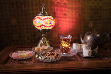 Arabian tea in glass with eastern snacks on vintage wooden surface. Eastern tea concept. Low light lounge interior with carpet. Empty space.
