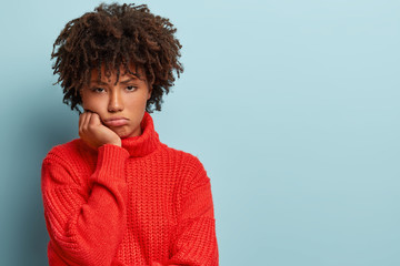 Human emotions concept. Displeased dark skinned woman with Afro haircut, gloomy expression, holds chin, feels sad and offended after quarrel with friends, wears red jumper, poses indoor alone