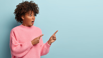 Indoor shot of impressed Afro American woman points aside with both fore fingers, dressed in pink jumper, has surprised facial expression, models over blue background. People, reaction, ethnicty