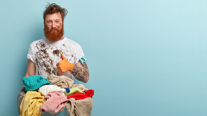 Self confident redhead man with beard keeps thumb up, shows like, did part of housework, has untidy clothes, stands in front of clothes basket with linen, stands over blue wall with free space