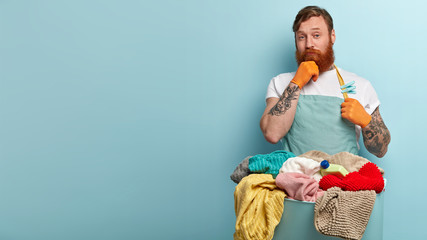 Indoor shot of curious red haired bearded busy man holds hand on chin, thinks which detergent to...