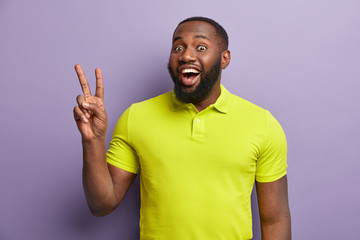 Glad unshaven black man with thick bristle, shows hand peace sign, likes something, wears bright...