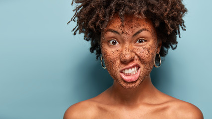 Displeased dark skinned woman frowns face with annoyance, doesnot want to make beauty procedures, has cleansing coffee mask on face, scrubs skin, stands over blue wall with empty space on left