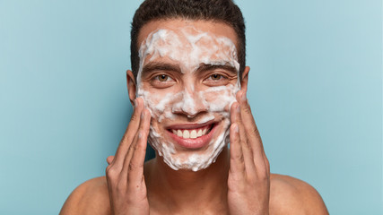 Photo of handsome satisfied European man cleans and washes face with soap, enjoys purity of skin,...