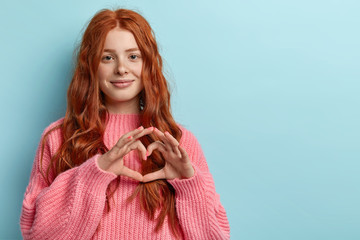 Portrait of beautiful red haired woman with freckled face, shows heart gesture over chest, wears...