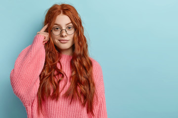 Photo of attractive ginger pupil with long wavy hair, tries to memorize something, keeps fore finger on temple, reminds information for exam, wears spectacles and jumper, stands over blue wall - obrazy, fototapety, plakaty