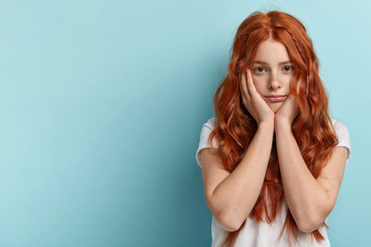 Indoor shot of sad lonely ginger girl touches cheeks with both hands, feels sad without friends, dressed in casual t shirt, has natural long curly red hair, freckled skin, models indoor over blue wall