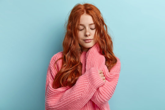 Photo of lovely ginger woman with freckled face, has gaze focused down, thinks about starting new relationships, wears loose knitted jumper, models over blue background. Red haired women are around us