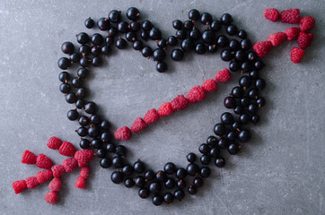 Fresh black currants in a shape of heart and a raspberry arrow on a grey table. Top view