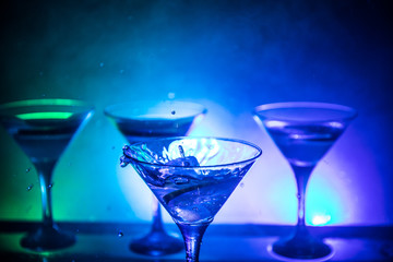Martini cocktail glass in hand splashing on dark toned smoky background or colorful cocktail in glass with splashes and olives. Party club entertainment. Mixed light. Selective focus
