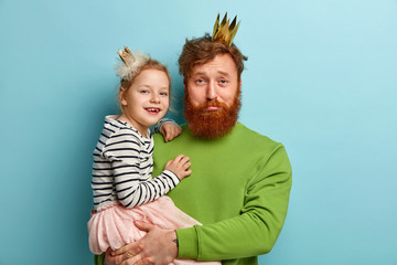 Funny family concept. Sad bearded ginger man and his happy child daughter wear crowns, prepare for...