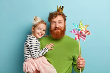 Happy ginger family of father and daughter wear party accessories, hold striped toy windmill,...