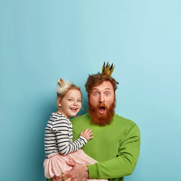 Vertical shot of surprised bearded red haired father carries small daughter, celebrates childs birthday, shocked to see many guests, have family celebrations. Parenthood and fatherhood concept