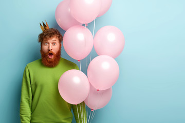 Shocked ginger dad keeps jaw dropped, recieves big present and balloons from children on Fathers Day, dressed in bright green jumper, has crown on head, celebrates festive event, isolated on blue