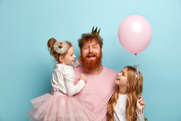 Isolated shot of upset gloomy man tired of naughty children, celebrates daughters birthday. Affectionate girls look at father, dressed in festive attire, hold air balloon isolated over blue background - obrazy, fototapety, plakaty