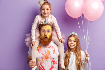 Surprised man entertains kids and organizes real holiday on Children day, being dirty with oil paints, piggybacks his little child lovely girl with crown holds air balloons. Party time and fun concept