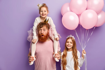 Emotional red haired man entertains children on party, surprised to do something wrong, carries small child on shouldres, positive kid with long hair holds balloons, congratulates friend with birthday