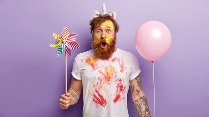 Indoor shot of surprised red man stares with shock, holds balloon and windmill in both hands, wears unicorn headband with horn and ears, smearked with colorful paints, entertains children on party - obrazy, fototapety, plakaty