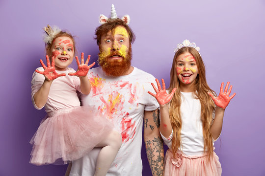 Funny stupefied dad and two adorable daughters show dirty palms with paints, stare at camera together, wear festive carnival costumes, celebrate Children Day, isolated over purple background