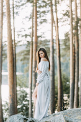 Girl in a boudoir dress in a pine forest. Standing on the rocks in a thin flying dress. Unity with nature at the wedding. Bride at sunset