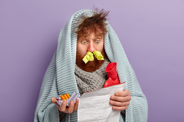 Healthcare concept. Unhealthy stupefied foxy man stares at medical leaflet, surprised with side effect of tablets, warms with coverlet and hot water bag, cures fever and cold, has tissues in nostrils