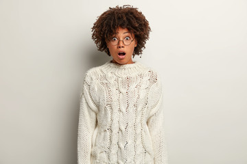 Monochrome shot of stupefied dark skinned woman with curly Afro hair, opened mouth, surprised to hear shocking news, wears white knitted jumper and spectacles, impressed by awful revelation.