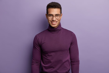 Satisfied European guy wears puple turtleneck sweater, round spectacles, feels pleasure and smiles, happy to come on meeting with colleague, isolated over lilac studio wall. Masculinity concept