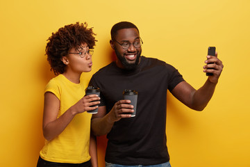 Lovely Afro woman keeps lips folded, poses at camera of cell phone for making selfie portrait together with boyfriend, drink takeaway coffee, creat publication, click images, isolated over yellow wall