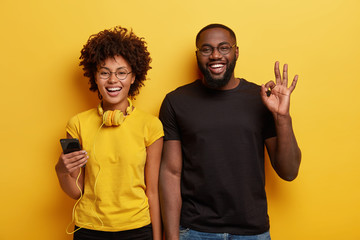 Waist up shot of joyful Afro American woman and man enjoy leisure time, listen music on cell phone. Dark skinned guy makes okay gesture, likes melody, stands near girlfriend. Lifestyle concept - obrazy, fototapety, plakaty